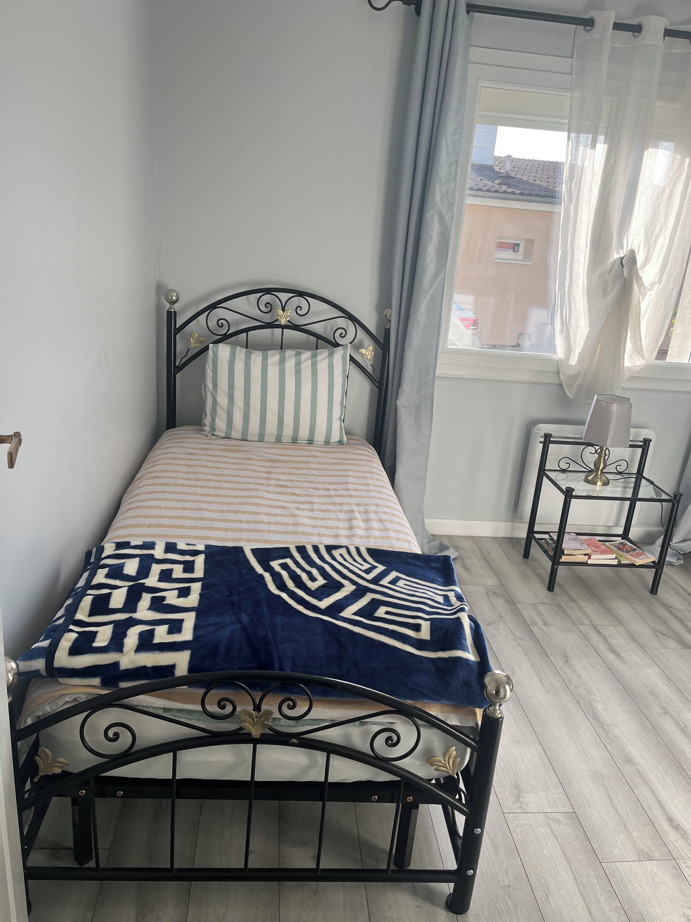 Un letto singolo con struttura in metallo, un cuscino a righe, una coperta con motivo blu e bianco e un comodino con dei libri.
