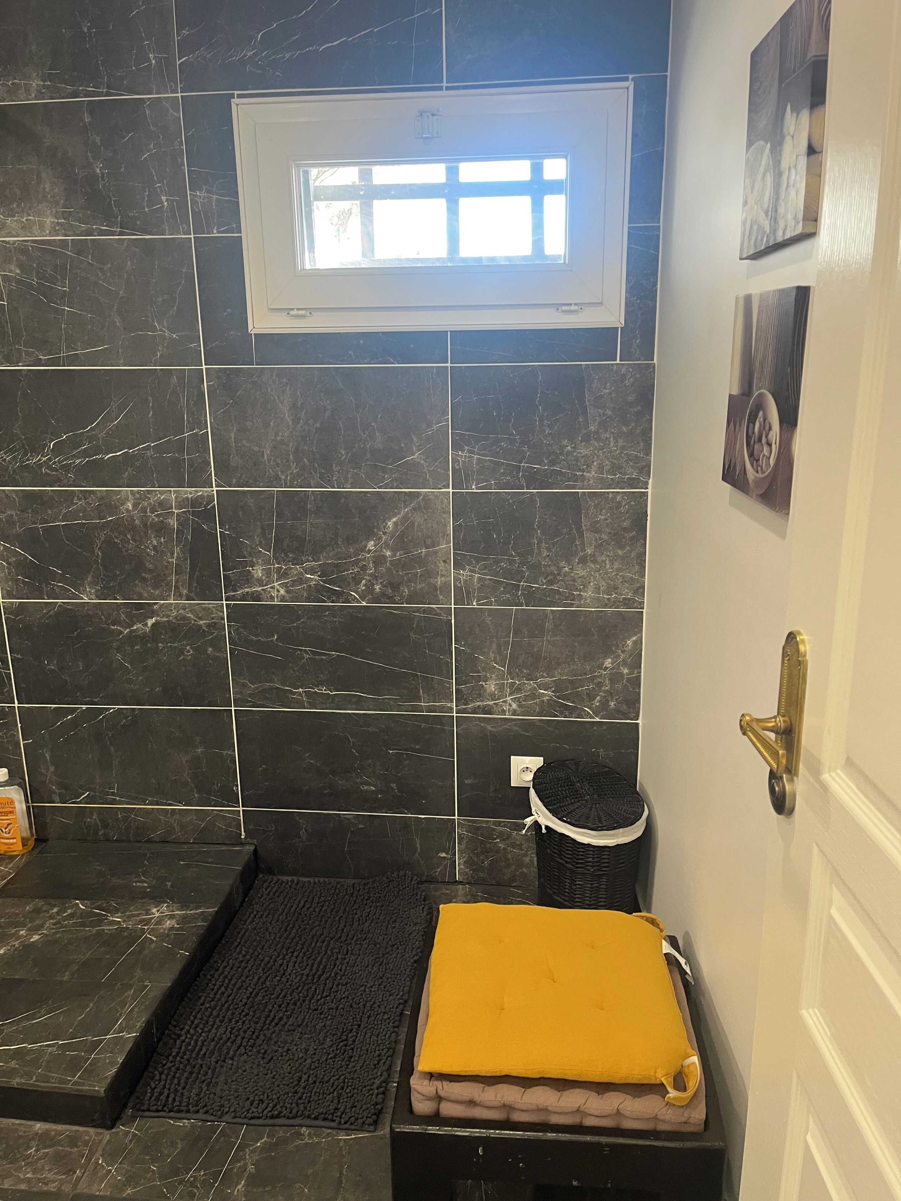 Une salle de bain avec des carreaux de marbre foncé, une petite fenêtre, un porte-serviettes et une porte dotée d’une poignée en laiton.