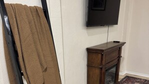 Ein Zimmer mit einem schwarzen Metallregal, auf dem eine braune Decke und ein weißes Handtuch liegen, einem Holzkasten mit einem Fernseher darüber und einem gemusterten Teppich auf dem Boden.