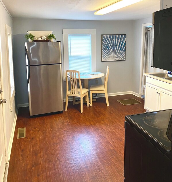 Una cocina compacta con refrigerador, una mesa de comedor con dos sillas, una ventana con persianas y un cuadro en la pared.