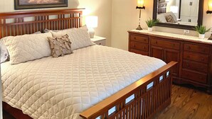 A bedroom with a large bed, wooden headboard, a ceiling fan, a nightstand with a lamp, a dresser, a mirror, and a framed painting on the wall.