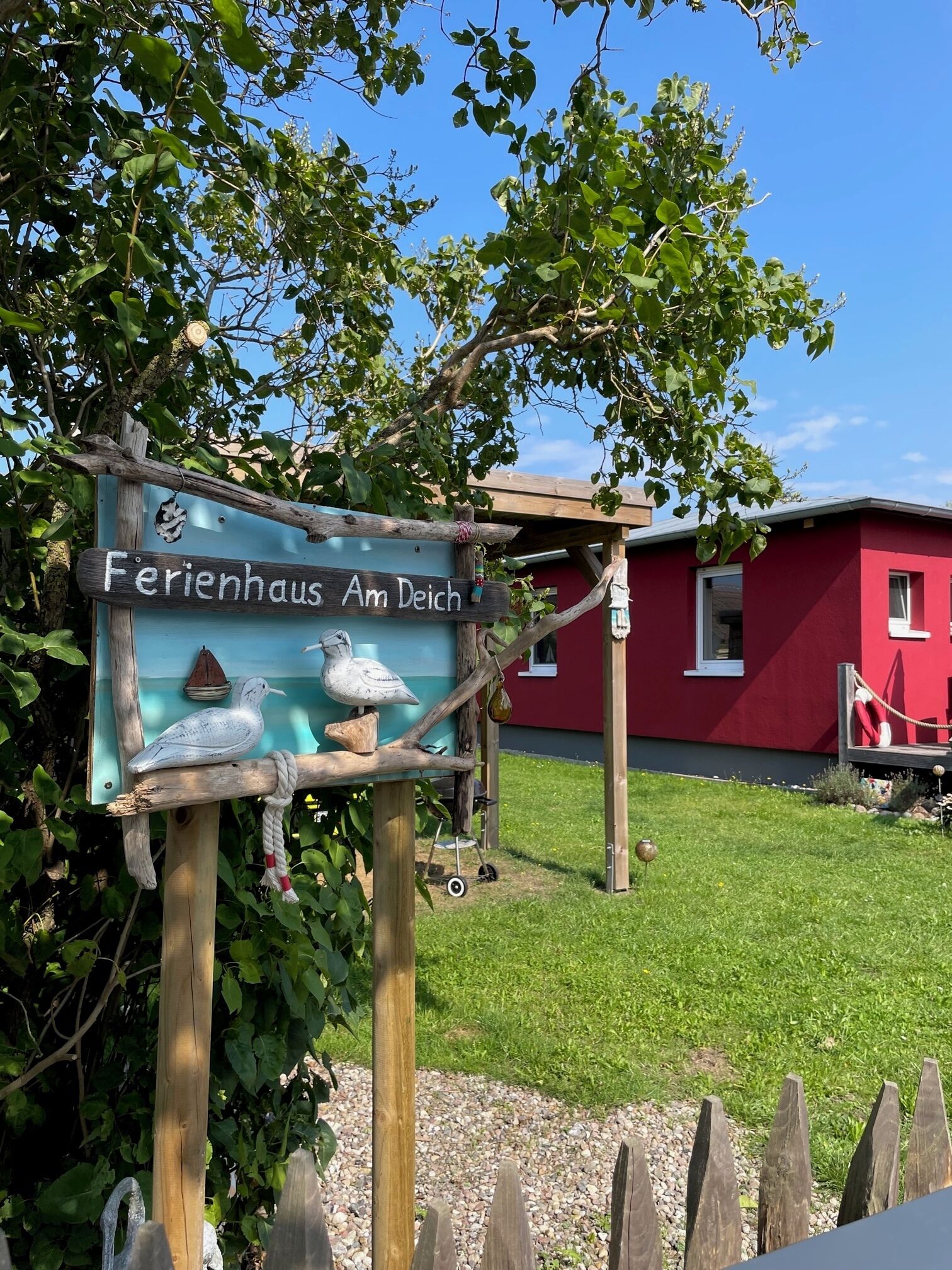 A sign for 'Ferienhaus Am Deich' with a wooden post, a bird sculpture, and a red building in the background.