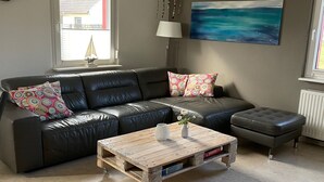 Ein Wohnzimmer mit einem schwarzen Ledersofa, einem Holztisch und Blick ins Freie durch ein Fenster.
