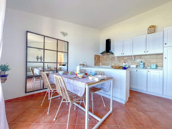 A compact kitchen with white cabinets, a tiled backsplash, and a dining area with wicker chairs and a table. - Oleander House, Costa Paradiso Li Cossi Hill (Costa Paradiso)
