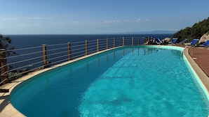 A clear blue pool with a wooden deck and lounge chairs, overlooking the sea.