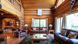 Interior de una cabaña de troncos con techo de madera, un sofá de cuero marrón, una mesa de centro de madera y un comedor visible al fondo.