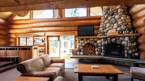 Interior de una cabaña de troncos con chimenea de piedra, sofás de cuero, mesas de madera y un televisor.