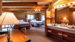 Una habitación tipo cabaña de troncos con interior de madera, una cama doble, un escritorio con lámpara, un espejo grande y un baño con lavabo y perchero para toallas.