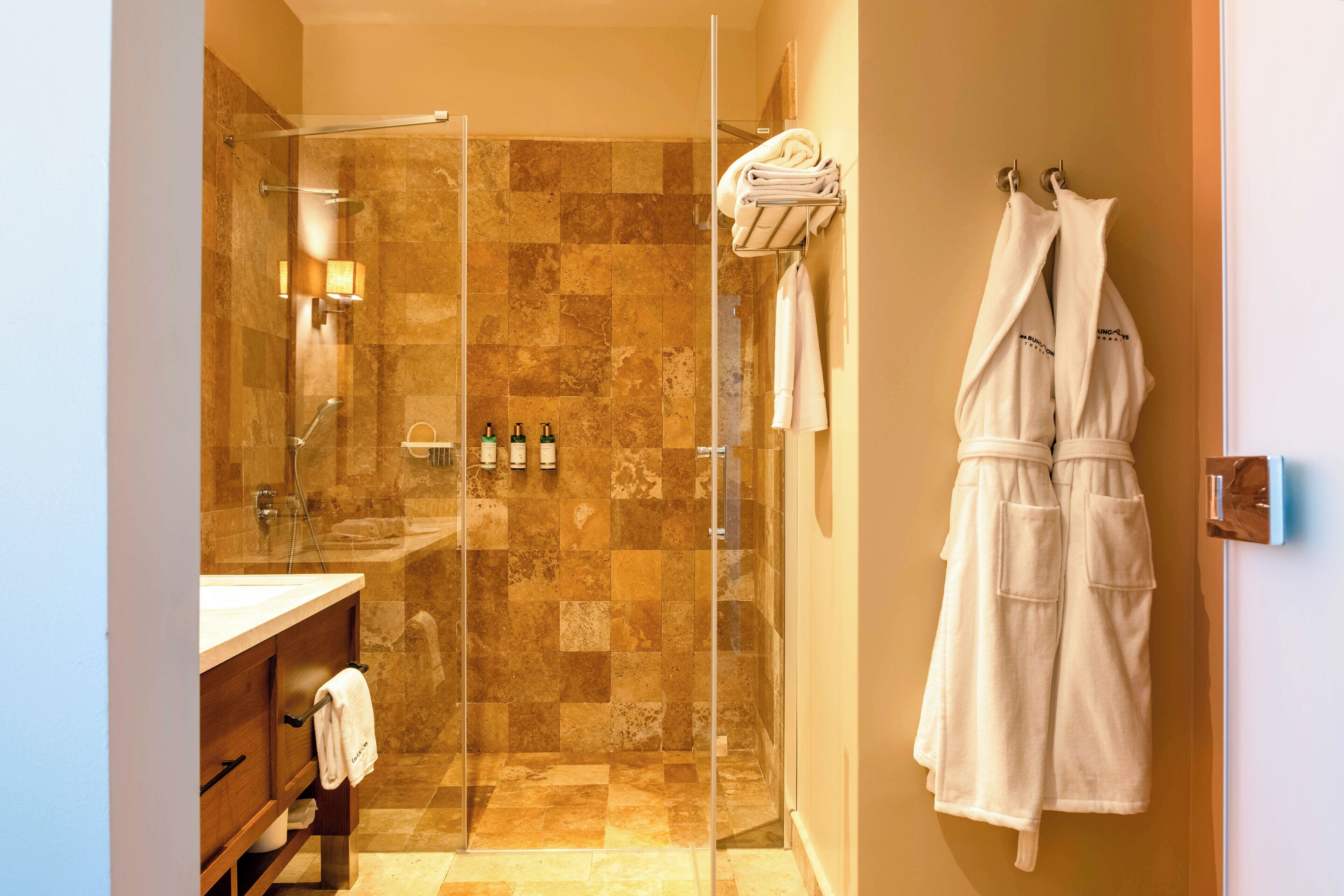 A hotel bathroom with a glass-enclosed shower, a vanity with a towel rack, and a robe hanging on a hook.