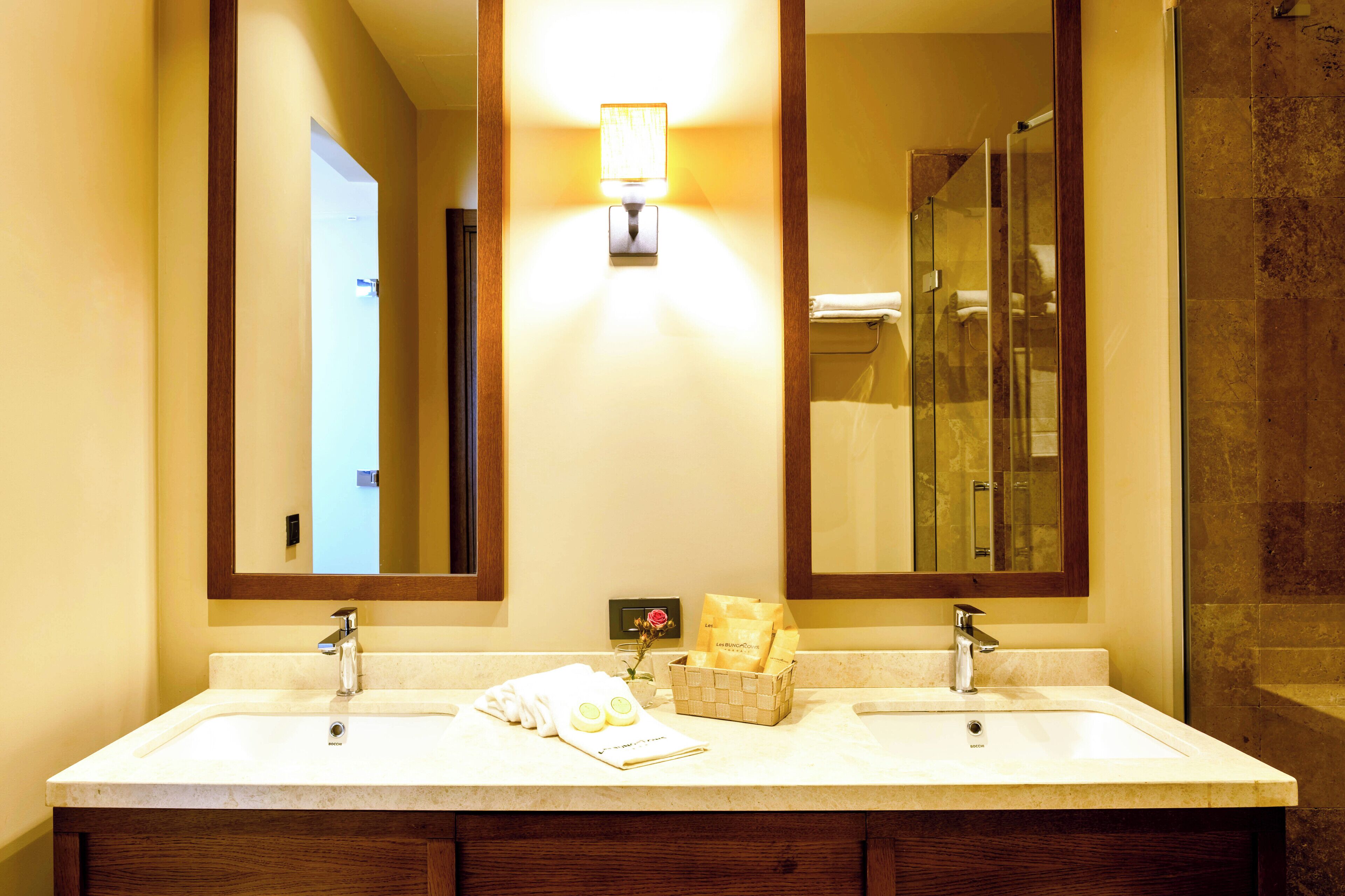 A hotel bathroom with two sinks, a mirror, a wall-mounted light, and a glass shower enclosure.
