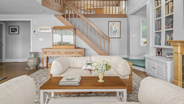 A living room with a wooden staircase, a white sofa, a coffee table with books, a vase with flowers, and a bookshelf.