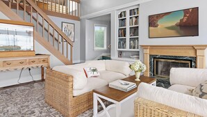 A spacious living room with a high ceiling, a wooden staircase, a fireplace, and a large bookshelf.