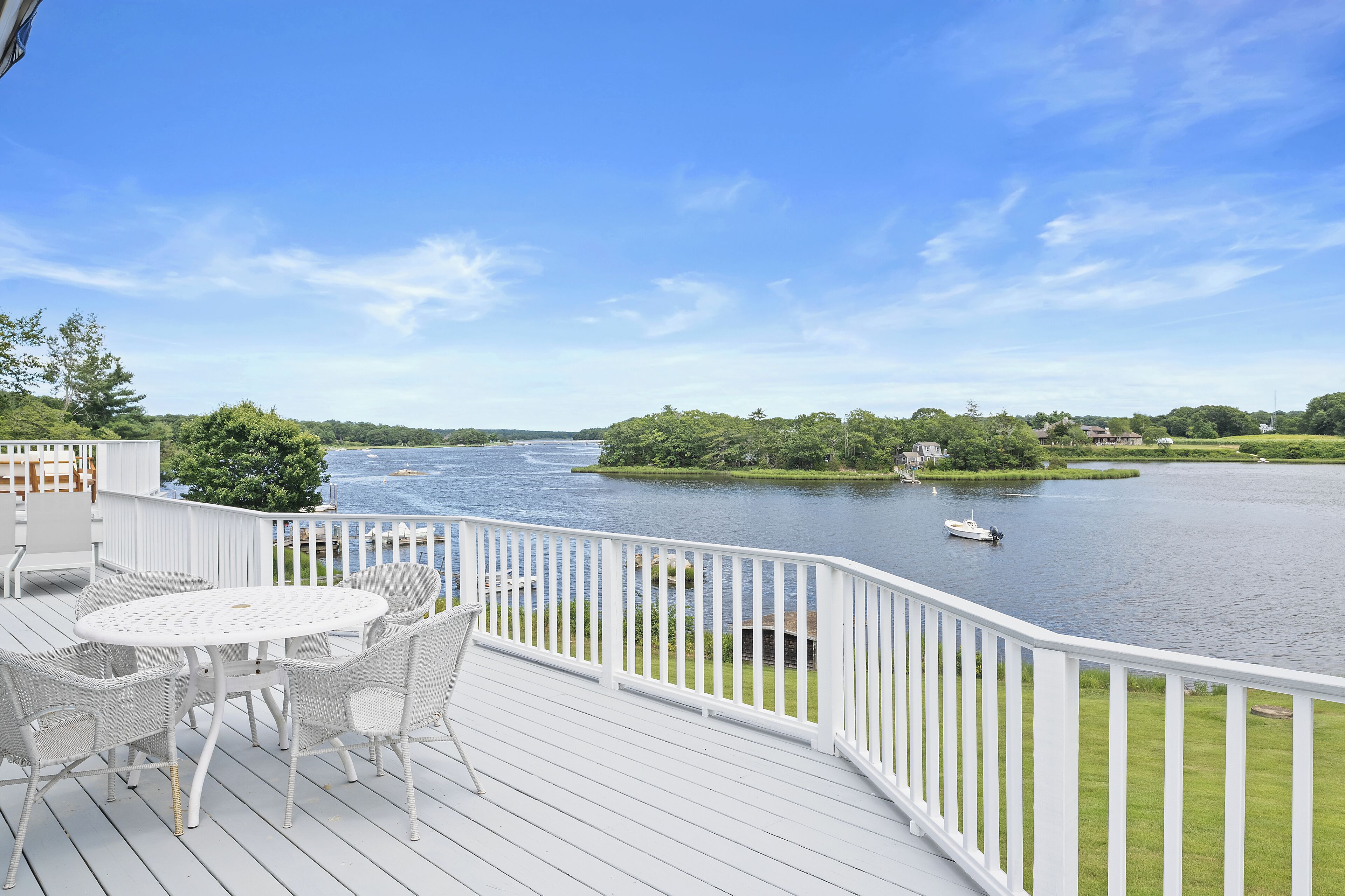 Une terrasse avec une table et des chaises blanches, donnant sur une rivière où se trouve un bateau.