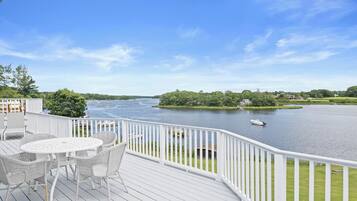 Une terrasse avec une table et des chaises blanches, donnant sur une rivière où se trouve un bateau.
