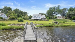 A wooden dock leading to a calm river, surrounded by lush greenery and large houses.