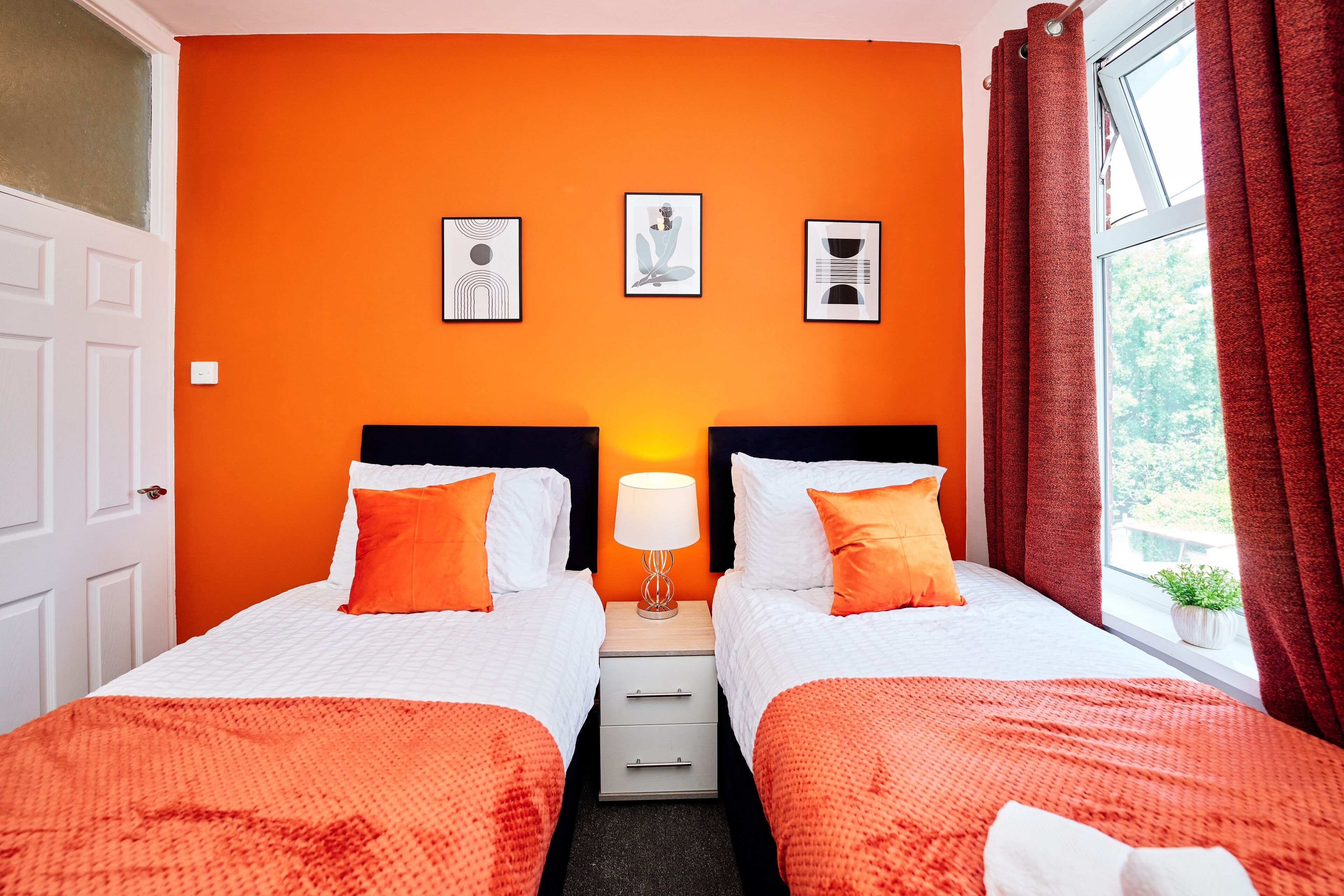 Ett hotellrum med två sängar, orange väggar, ett fönster med röda gardiner och tre inramade tavlor på väggen.