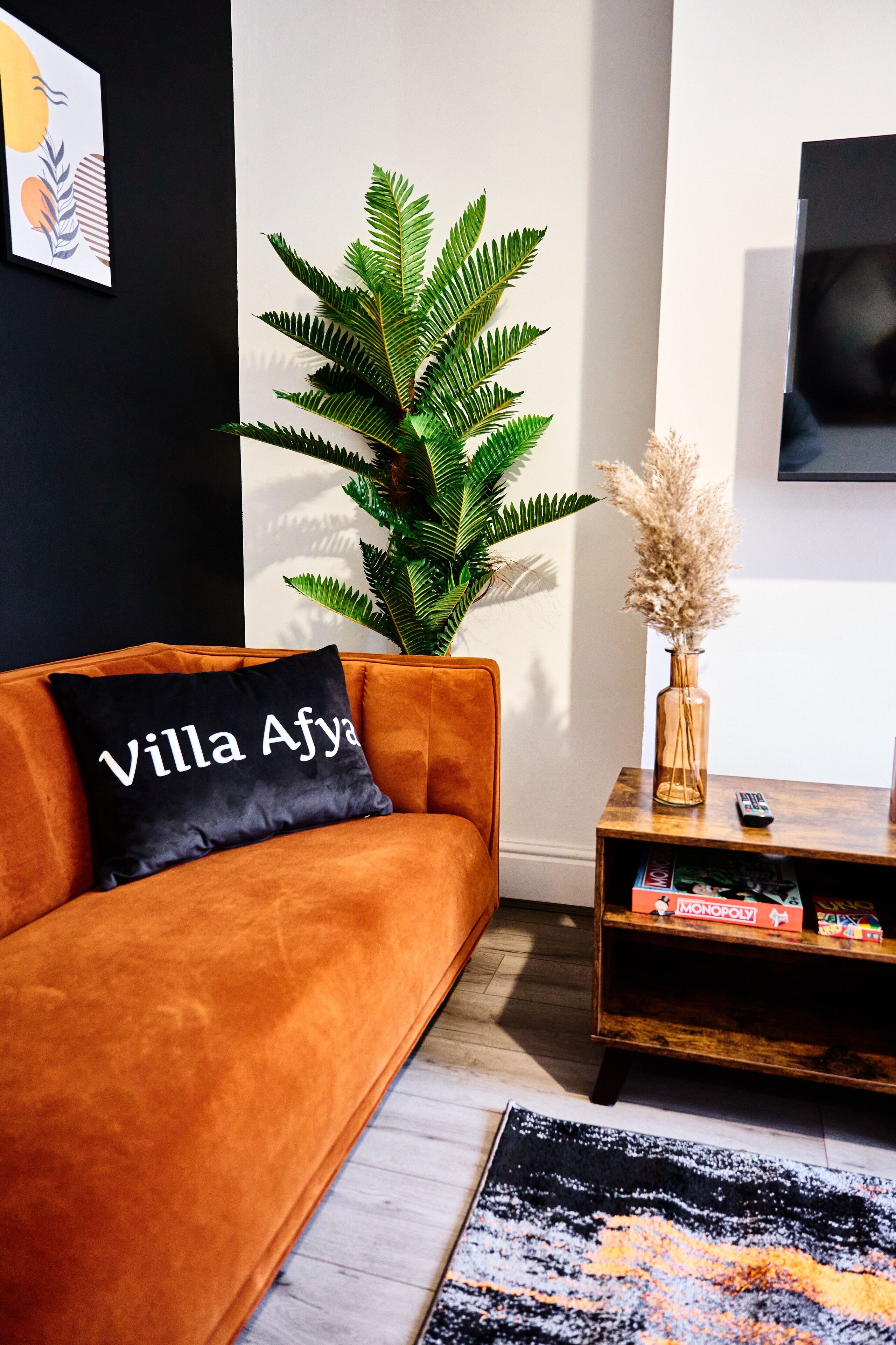 A modern living room with a sofa, a potted plant, a vase with dried flowers, and a wooden side table with a remote control.
