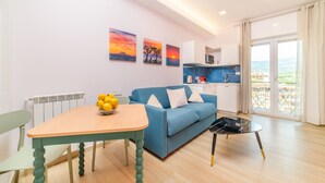 A modern living room with a blue sofa, a wooden dining table with a bowl of lemons, and a small black coffee table. There are three paintings on the wall depicting sunsets. - Domus Sirenum - Teles (Sorrento)