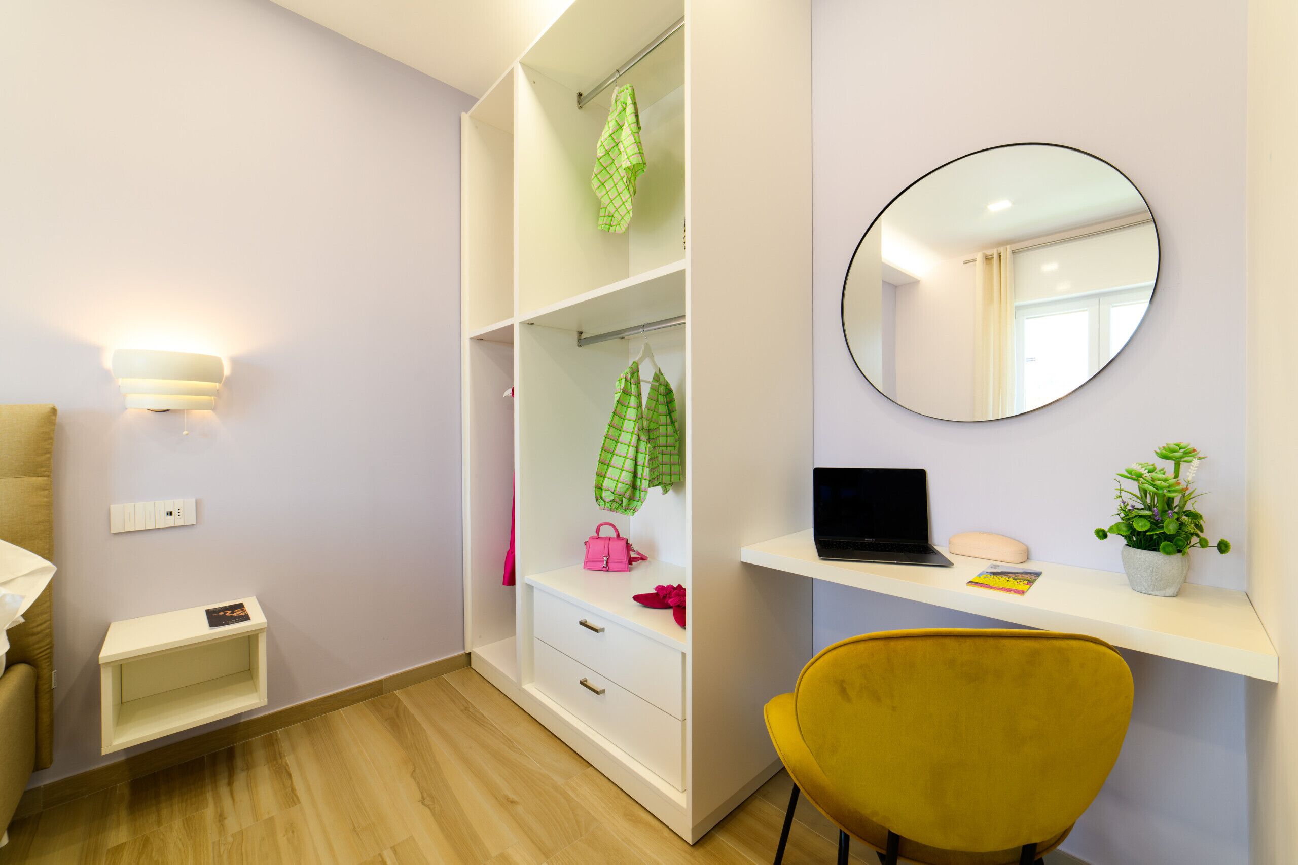 Une chambre moderne équipée d’un lit, d’un bureau avec un ordinateur portable, d’un miroir et d’une armoire où sont suspendus des vêtements.