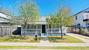 A two-story residential building with a front porch, a garage, and a small front yard with trees.