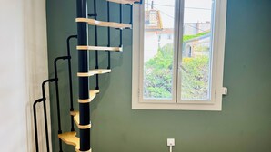 A room with a spiral staircase, a window with a view of greenery, and a light-colored wooden floor.