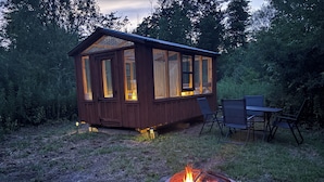Eine Holzhütte mit großen Fenstern, einer Feuerstelle und einem Esstisch mit Stühlen in bewaldeter Landschaft.