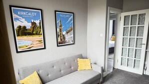 A modern living room with a grey sofa, two yellow cushions, and a wooden coffee table. There are two framed pictures on the wall depicting Kirkcaldy and Falkland.