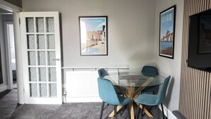 A dining area with a glass table and four blue chairs, a white door with frosted glass panels, and two framed pictures on the wall.