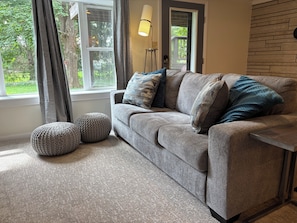 Un salon avec un canapé gris, deux poufs ronds, une lampe sur pied et une vue sur la verdure à l’extérieur.