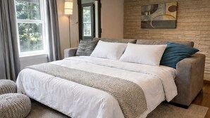 Une chambre avec un grand lit, un canapé gris, un plaid en tissu tissé et un mur en pierre orné d’un tableau.