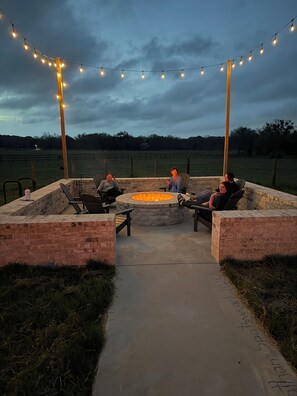 Un espace extérieur aménagé avec des sièges, un foyer et entouré de murs en briques et de guirlandes lumineuses.