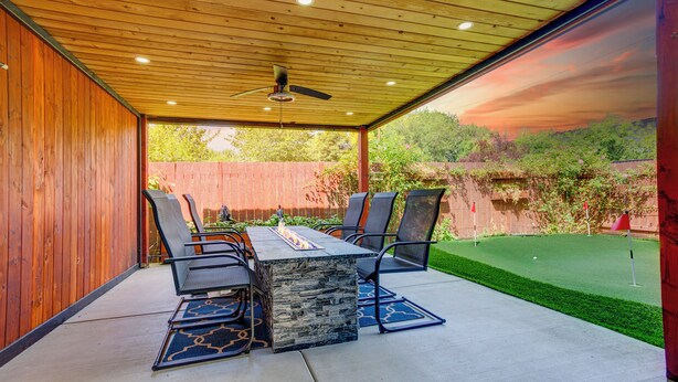 Un patio al aire libre con techo de madera, una mesa con sillas y vista a un césped verde y árboles.