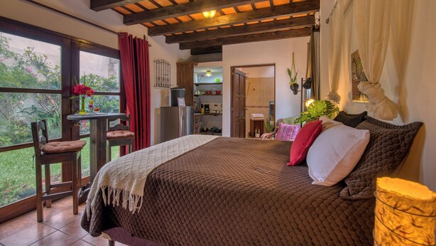 Un dormitorio con una cama grande, techo de madera, ventana con cortinas rojas y vista a la vegetación exterior.