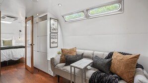 Un dormitorio moderno con un sofá gris, una mesita y un armario. Dos ventanas ofrecen vistas a zonas verdes.