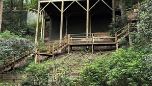 Une cabane en bois dotée d’un balcon, entourée d’une végétation luxuriante et d’un mur de pierres.