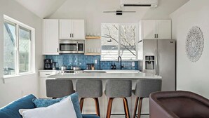 A modern kitchen with white cabinets, a central island, bar stools, and a refrigerator. - E ATX Tree House | Deck | Fire Pit | BBQ | Parking (Austin)