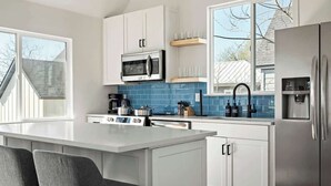 A modern kitchen with white cabinets, a built-in microwave, a stainless steel refrigerator, and a large island with bar stools. - E ATX Tree House | Deck | Fire Pit | BBQ | Parking (Austin)