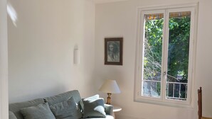 Una sala de estar con un sofá gris, un tapete estampado, una mesa de centro de vidrio, una lámpara y una ventana con vistas a la vegetación.