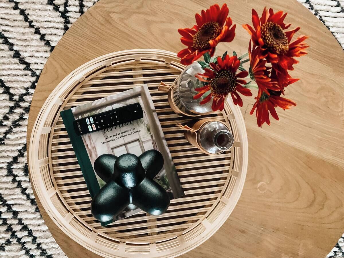 A round wooden table with a woven tray, a book, decorative stones, and red flowers.