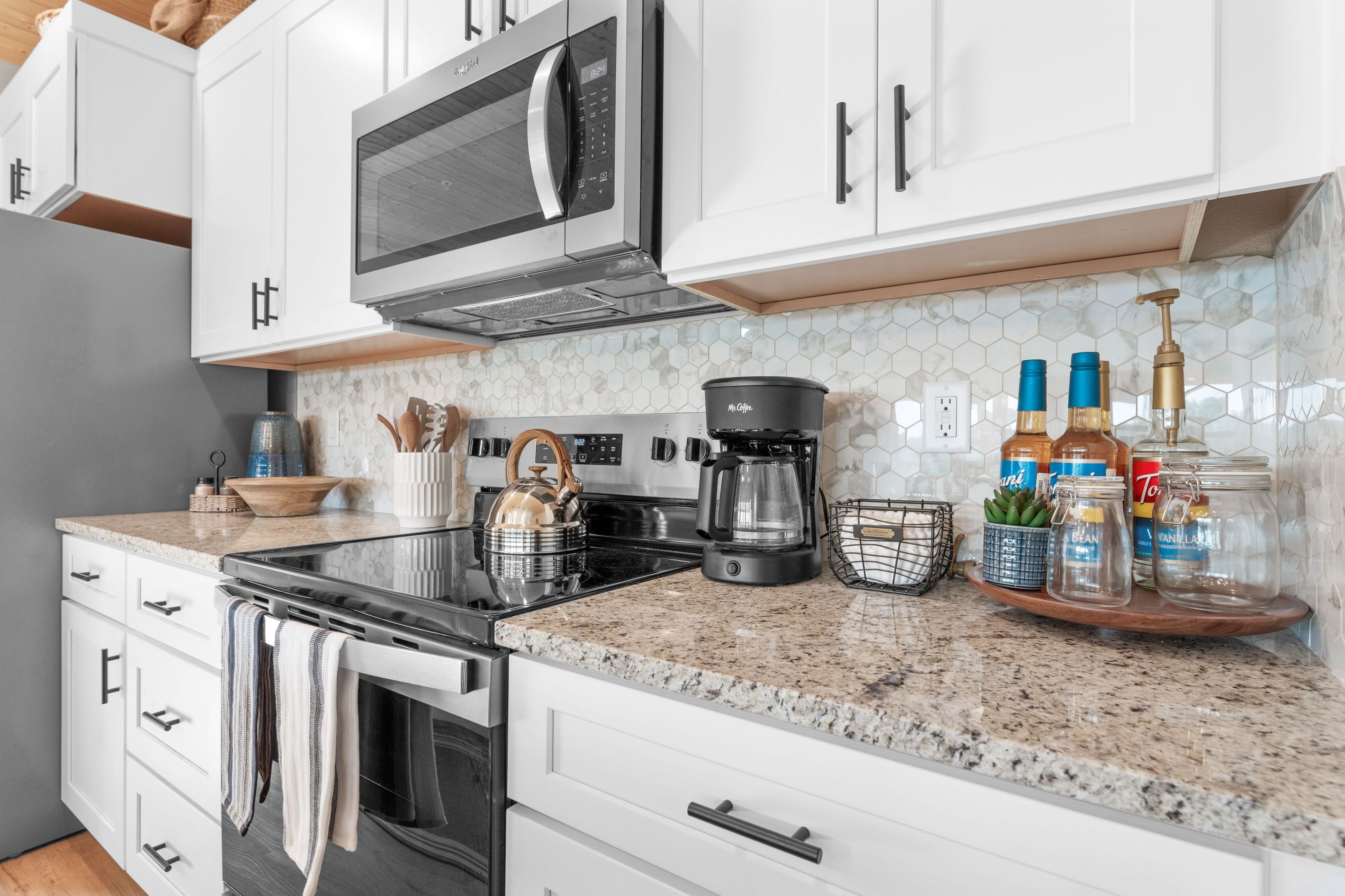 Una cocina moderna con armarios blancos, microondas, cafetera y diversos utensilios de cocina.