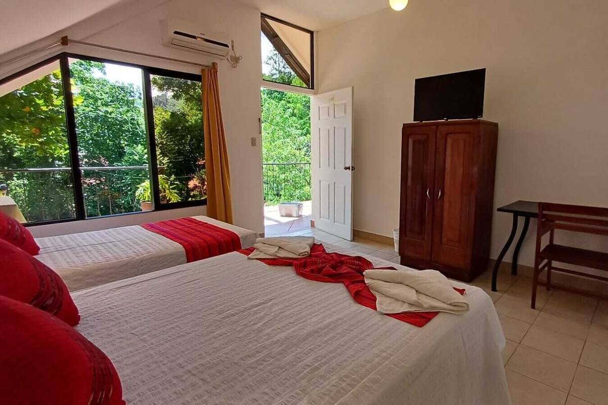 Une chambre d’hôtel avec deux lits, une armoire en bois, une télévision, un banc et une vue sur la verdure par la fenêtre.