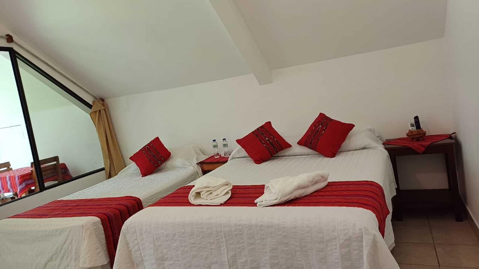 Une chambre d’hôtel avec deux lits, une literie rouge et blanche, une petite table avec un téléphone et des bouteilles d’eau, et une fenêtre donnant sur un paysage agréable.