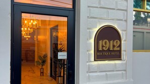 L’entrée d’un hôtel, avec une porte noire, une enseigne indiquant « ENTRÉE » et une plaque portant l’inscription « HÔTEL BOUTIQUE 1912 ».