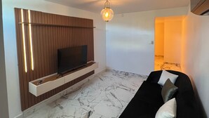 Un salon moderne avec un téléviseur à écran plat fixé sur un mur recouvert de panneaux en bois, un canapé noir avec des coussins et un sol à motifs de marbre.