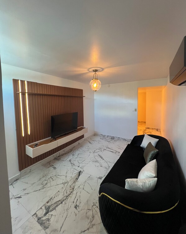 A modern living room with a flat-screen TV mounted on a wooden panel wall, a black sofa with cushions, and a marble-patterned floor.