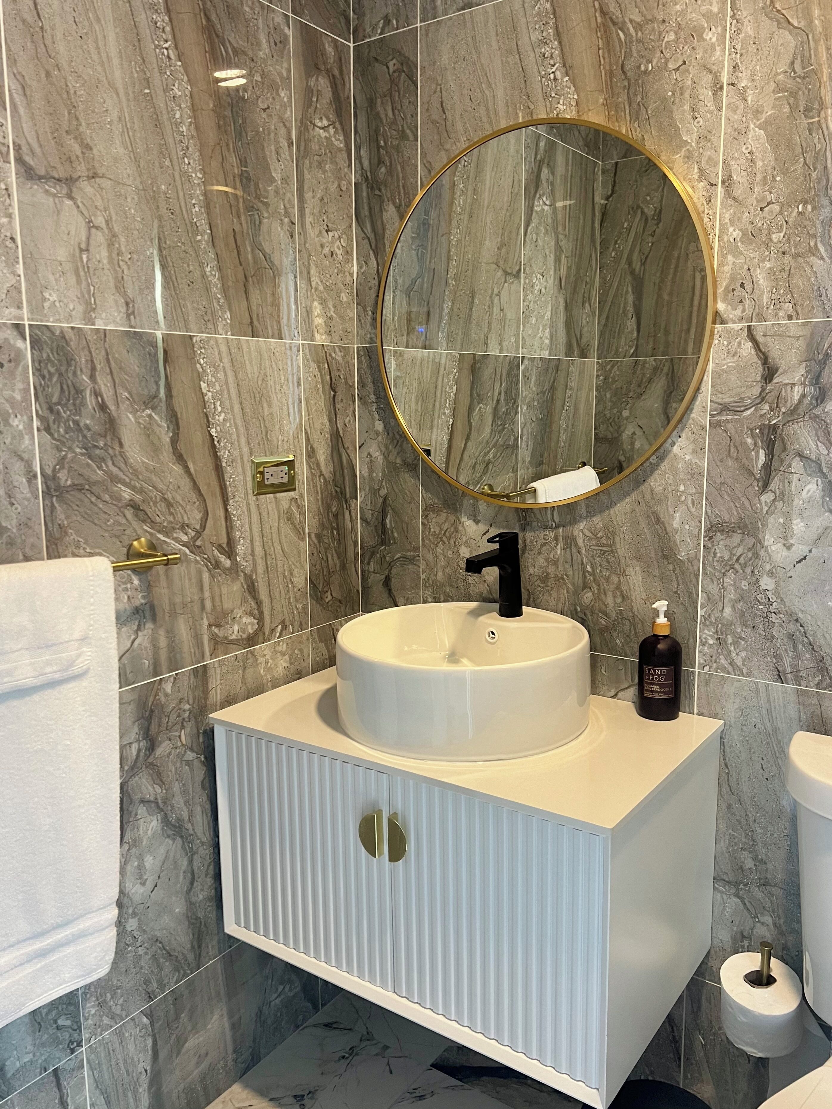 A bathroom with a marble wall, a round mirror, a white sink, and a toilet.
