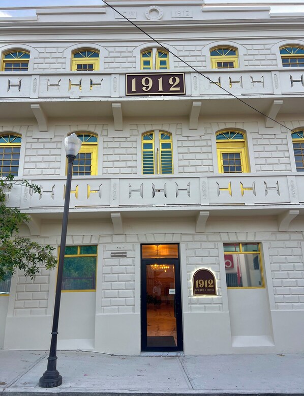 Un edificio con un llamativo letrero que dice '1912', varias ventanas con contraventanas amarillas y una puerta con un letrero que indica '1912 BOUTIQUE HOTEL'.