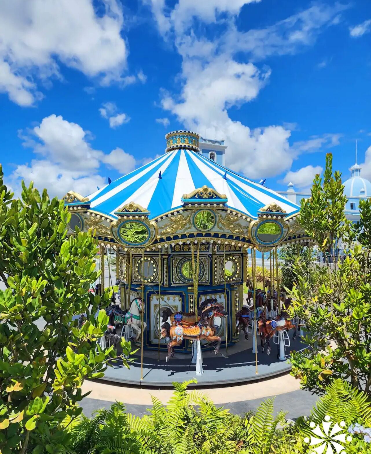 Un carrousel agrémenté de décorations dorées et de toits rayés de bleu et de blanc, entouré de verdure.