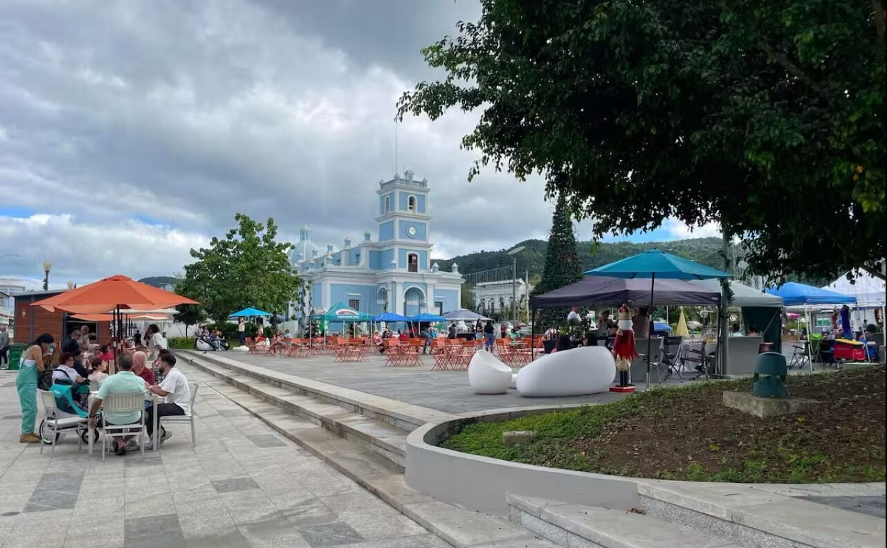 Une place publique avec, en arrière-plan, un bâtiment imposant aux couleurs bleu et blanc, entourée d’arbres et agrémentée d’espaces de repos en plein air.