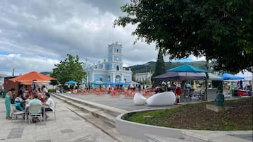 Ein öffentlicher Platz mit einem markanten blau-weißen Gebäude im Hintergrund, umgeben von Bäumen und Sitzgelegenheiten im Freien.
